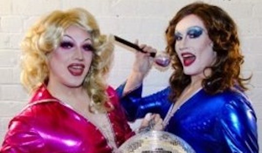 Two drag queens hold a microphone suggestively behind a big disco ball.