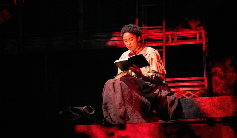 Mina sits reading a book, another on her lap. She is surrounded by red light. 