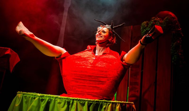 The performer, Ruxy Cantor, sits on a green table, legs akimbo, dressed as a tomato.